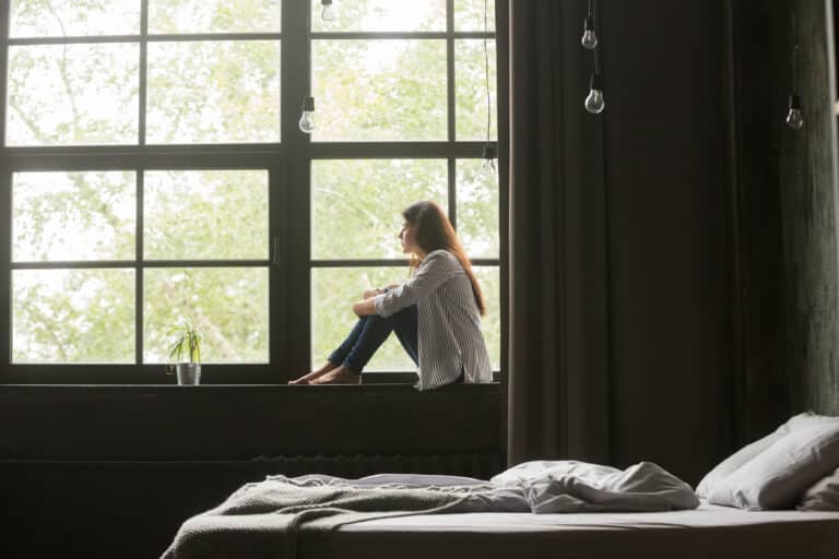 Lonely and depressed young woman sitting on windowsill at home
