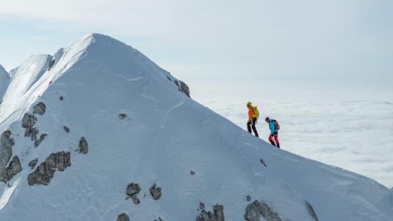 Two experienced mountaineers are walking on a snowy mountain ridge, equipped with winter climbing gear. They are navigating the icy terrain under clear skies, demonstrating teamwork and resilience.