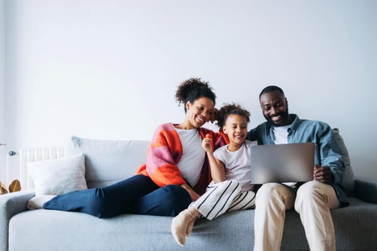 Happy family sitting on the couch using technology and practicing good digital wellness and learning more about online safety.