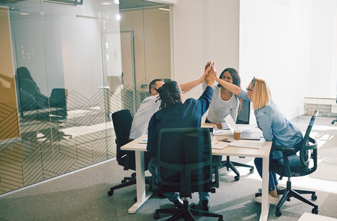 Diverse group of work colleagues smiling and high fiving together during a meeting around a table in a bright modern office while having a positive experience