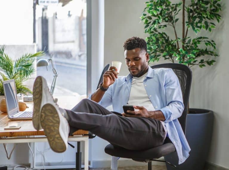 Man relaxing with coffee and his phone while procrastinating at work